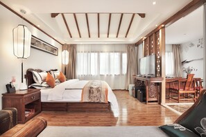 Quarto de hotel com cama, mesa de cabeceira, abajur, cortinas, uma escrivaninha com televisão e área de refeições com cadeiras e mesa.
