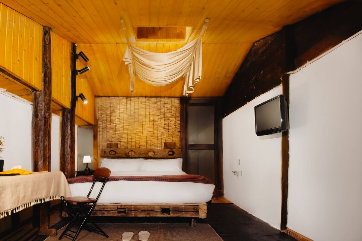 Een knus slaapkamer met een houten plafond, een bed met een geweven hoofdbord, een stoel, een nachtkastje en een tv aan de muur.