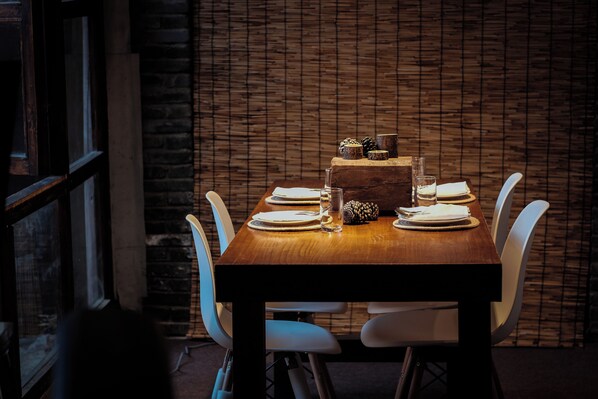 A wooden dining table set with white chairs, plates, and glassware against a brick wall.