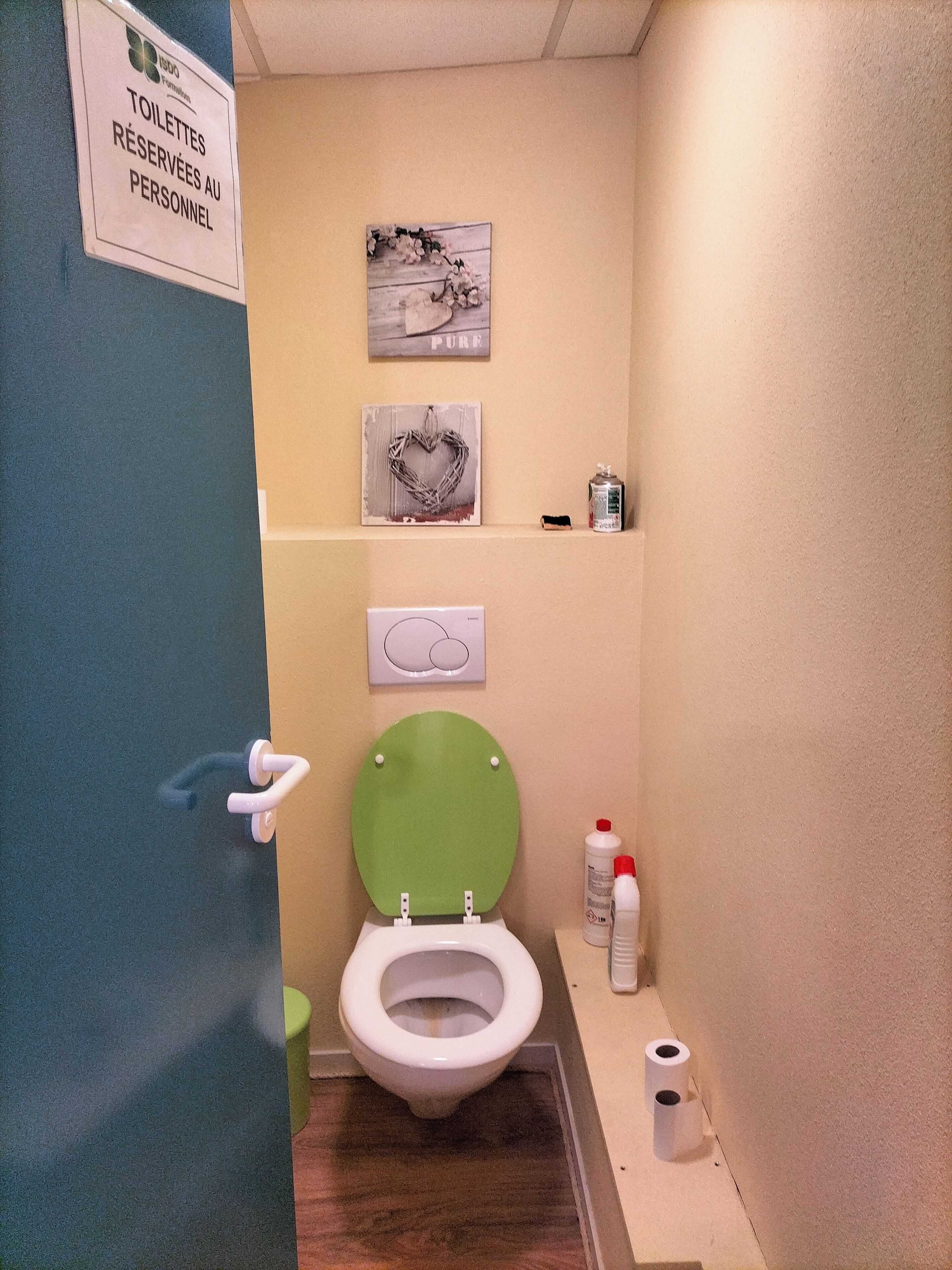 A small, private bathroom with a green toilet seat, a white toilet, and a sign indicating it is reserved for staff. There are two framed pictures on the wall, a toilet paper dispenser, and a small shelf with a bottle.