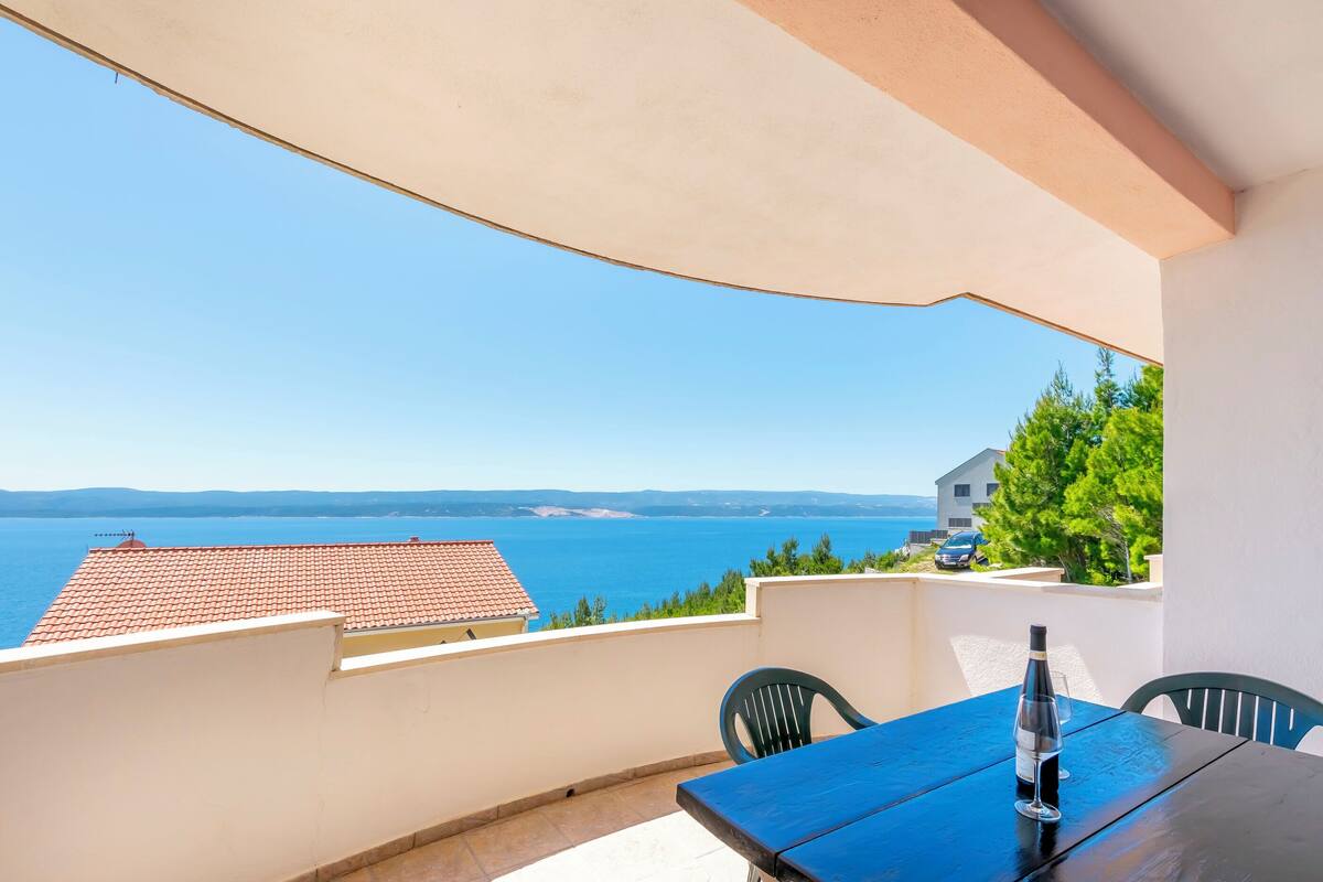 A balcony with a table and chairs, overlooking a coastal view with a clear sky.