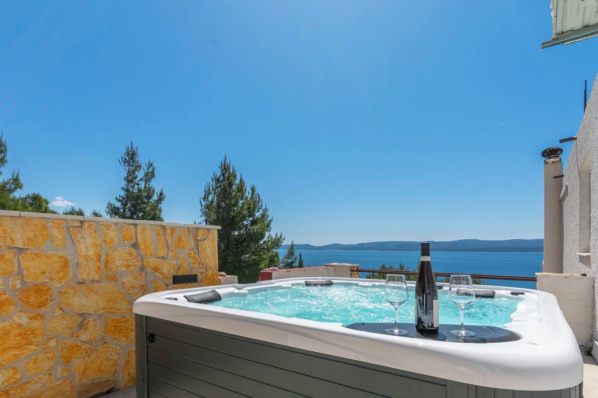 A hot tub with a bottle and glass, overlooking a body of water and mountains.