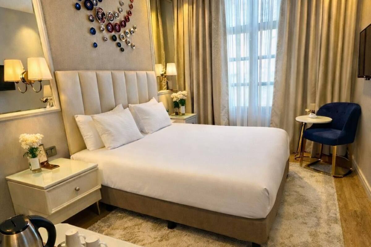 Une chambre d’hôtel avec un grand lit, des tables de chevet avec des lampes, un bureau avec une bouilloire et des bouteilles d’eau, et une chaise.