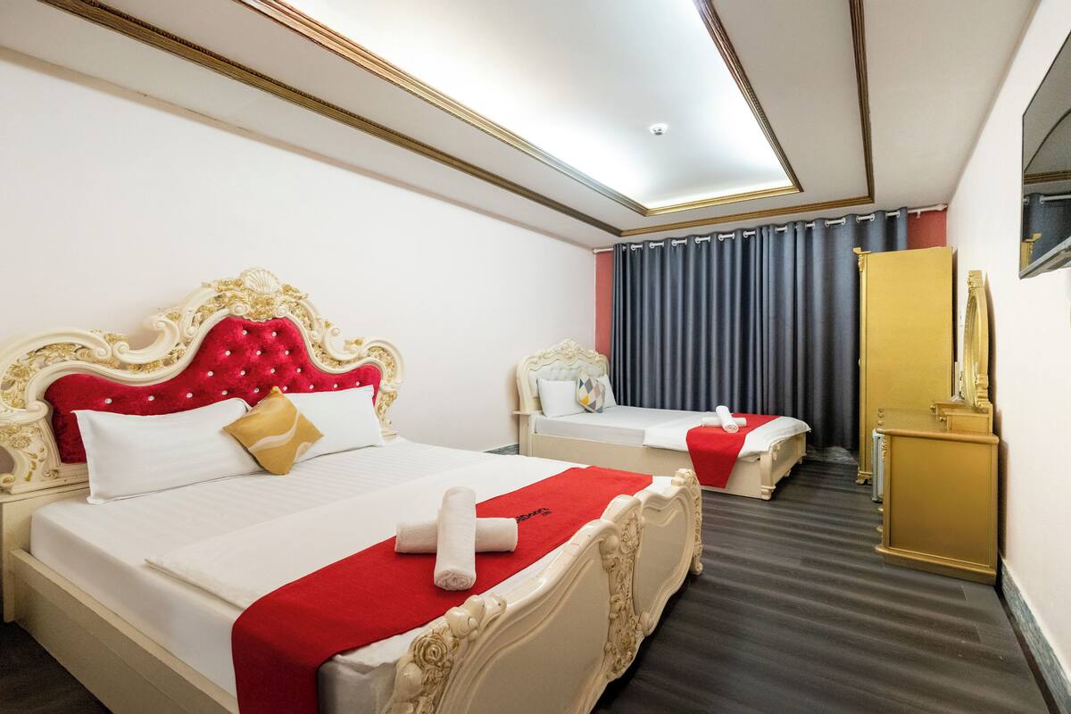 Habitación de hotel con dos camas, cada una con cabeceros ornamentados y detalles en rojo, una mesita de noche pequeña y un espejo en la pared.