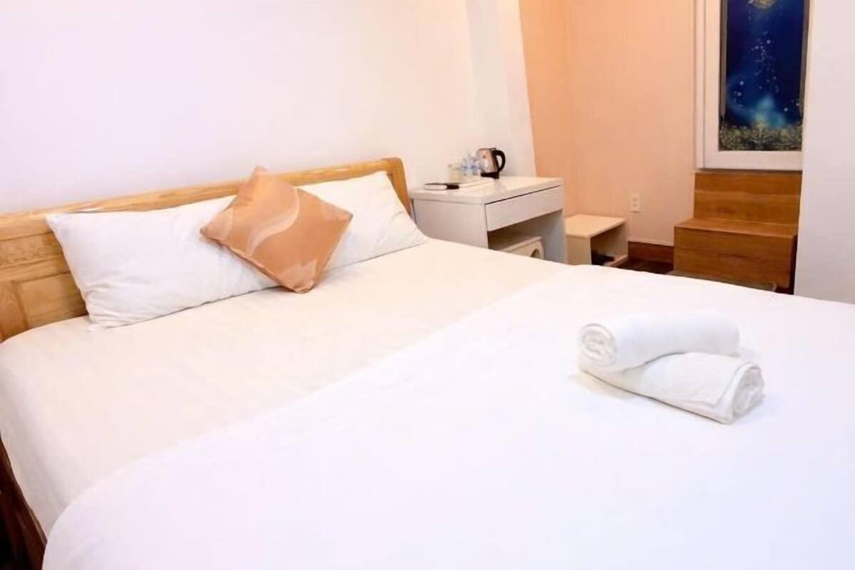 Una cama bien hecha con ropa de cama blanca y beige, una mesita de noche con una taza y un libro, y un cuadro en la pared.