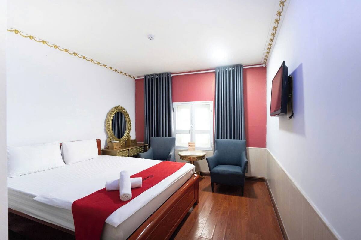 Una habitación de hotel con una cama, dos sillones, una mesa redonda, un espejo y un televisor en la pared.