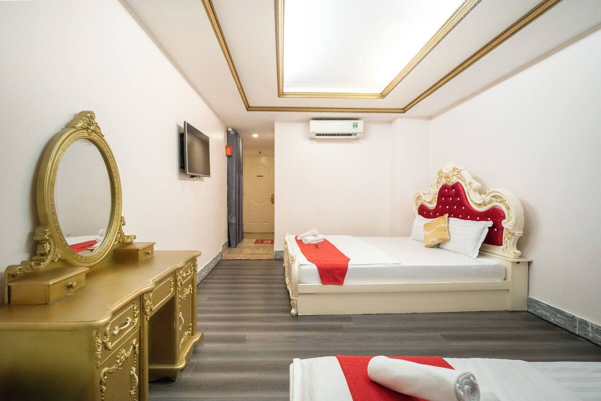 Una habitación de hotel con un espejo grande y ornamentado, una cama con cabecera roja y blanca, y un televisor de pantalla plana montado en la pared.
