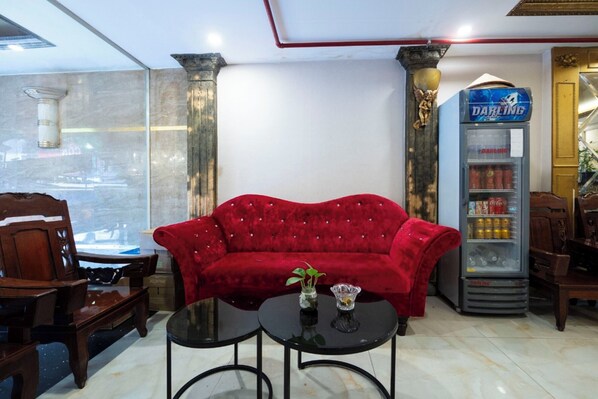 A red velvet sofa, two black tables, a vending machine, and wooden furniture in a room with marble flooring and a large window.