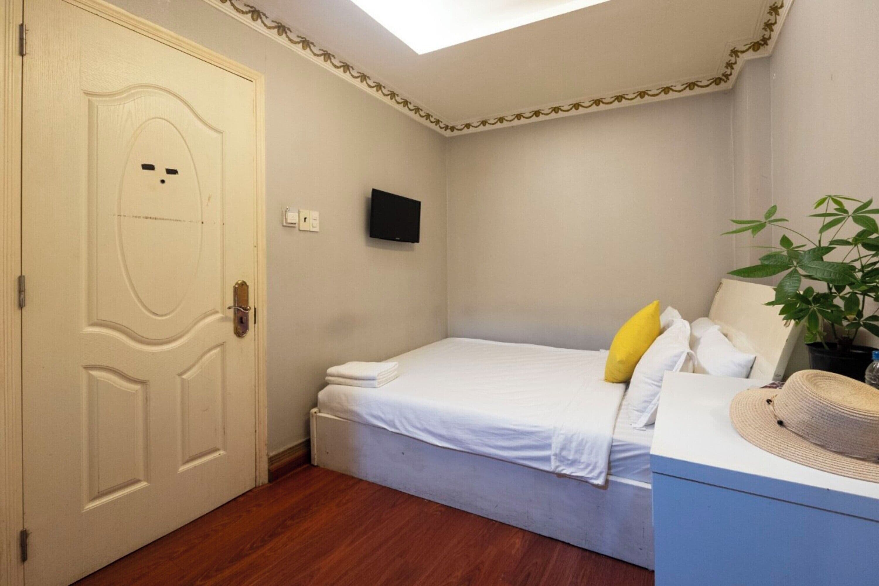 A bedroom with a single bed, a TV mounted on the wall, a plant, and a hat on a dresser.