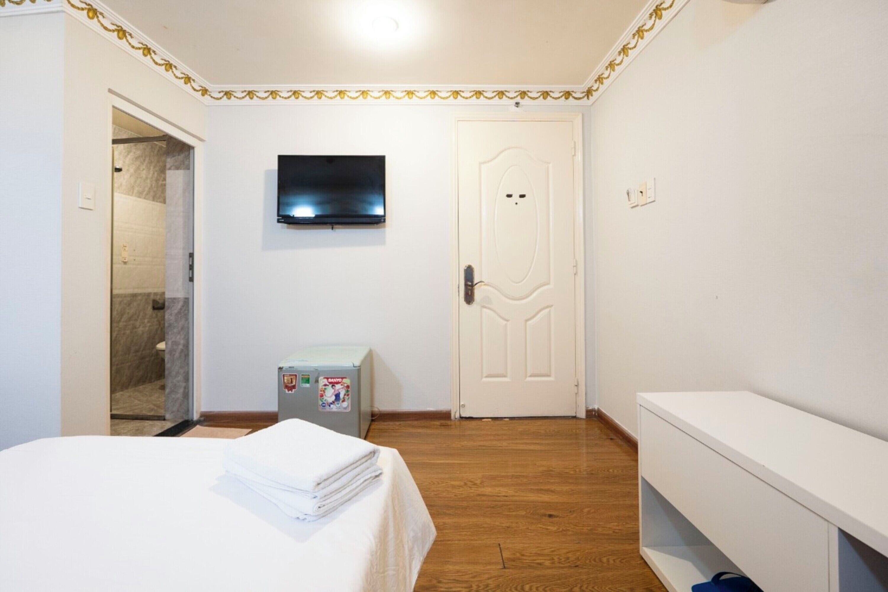 Un dormitorio con cama, televisión en la pared, puerta, un mueble pequeño y un banco.