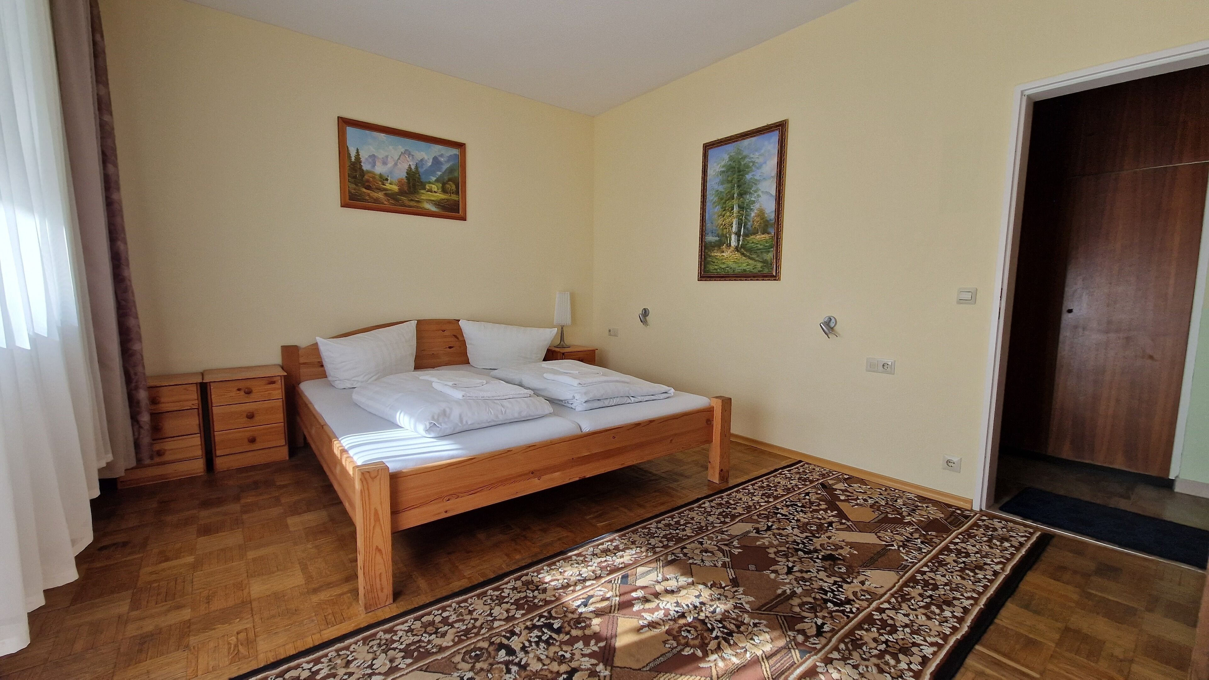 A bedroom with a wooden bed, two nightstands, a patterned rug, and two landscape paintings on the walls.