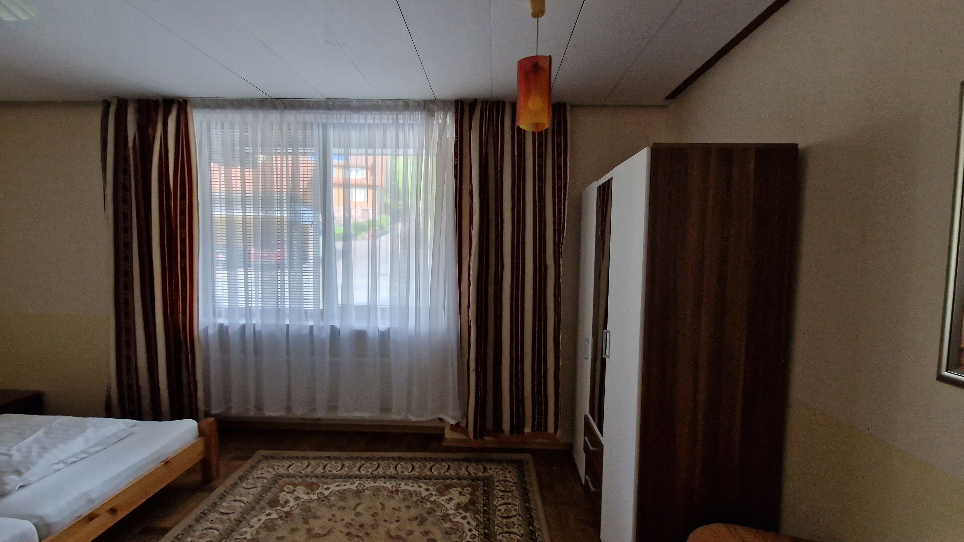 Un dormitorio con cama, armario de madera, alfombra estampada y ventana con cortinas.