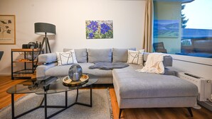 A modern living room with a grey sectional sofa, a glass coffee table, a black floor lamp, and a painting of blue flowers on the wall. - Domizil Deluxe Eleven (Kaprun)