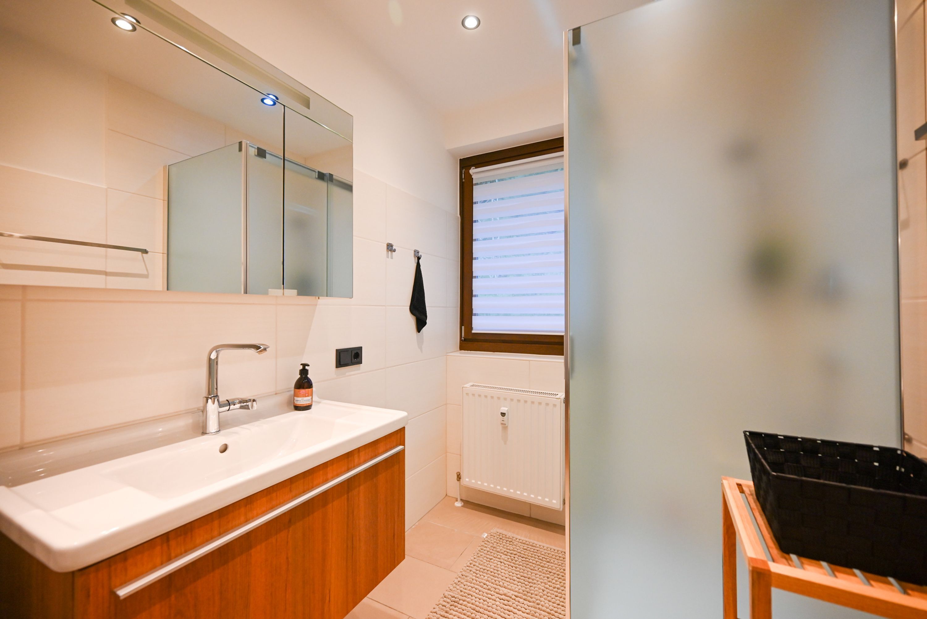 A modern bathroom with a white sink, wooden vanity, a mirror, a towel rack, and a radiator.