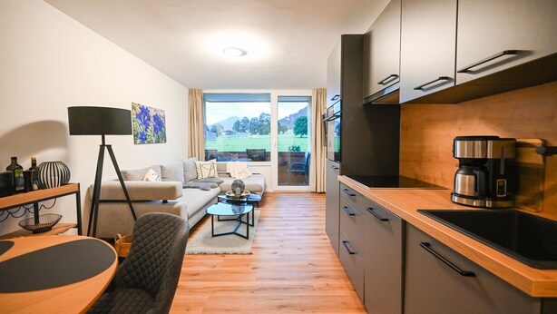A modern living room with a sofa, coffee table, and dining area. There is a kitchen with a coffee machine and cabinets. The room has a large window with a view of a green landscape. - Domizil Deluxe Eleven (Kaprun)