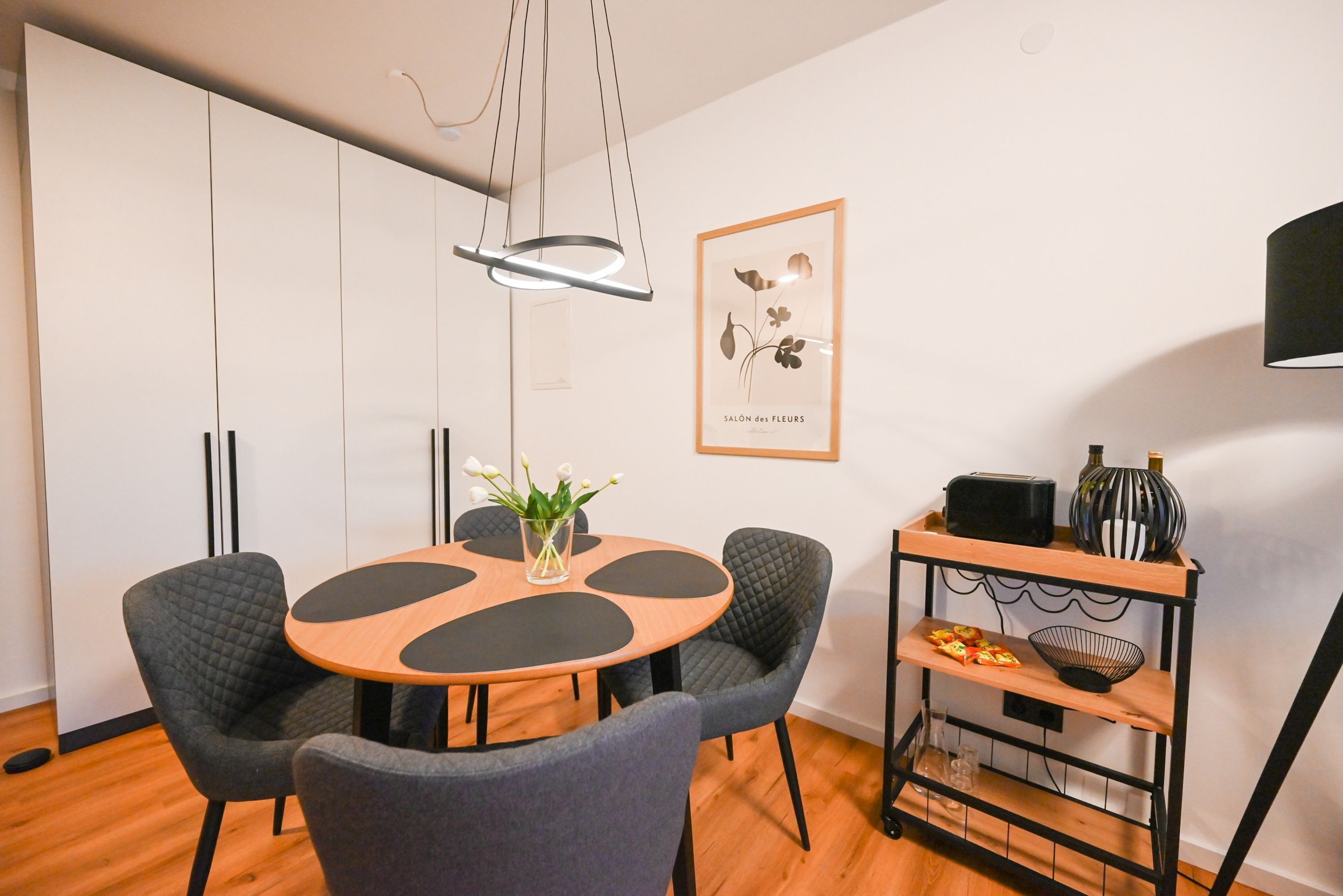 A modern dining area with a round table, four chairs, a pendant light, a framed botanical print, and a small side table with decorative items.