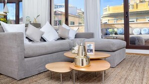 A modern living room with a grey sectional sofa, two round wooden side tables, and a large window offering a view of buildings. - Solaga Souviron Suits (Málaga)