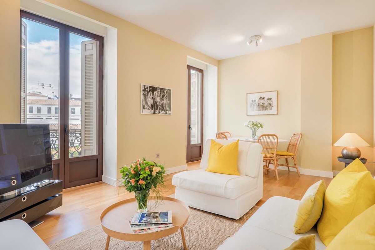 A modern living room with a flat-screen TV, a white sofa with yellow cushions, a round coffee table with magazines and a vase of flowers, and a dining area with wicker chairs.