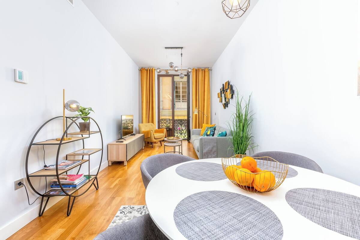 A modern living room with a round dining table, grey sofas, and a bookshelf.
