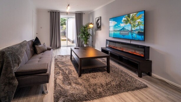 A modern living room with a sofa, a coffee table, a flat-screen TV mounted on the wall, and a fireplace. - Spacious 3 Bed House, 5mins to HS2/ NEC/Airport. (Birmingham)