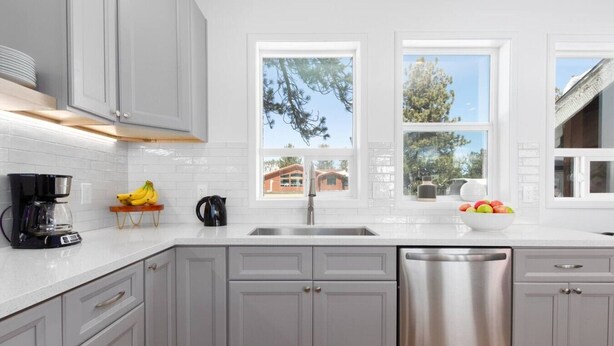 A modern kitchen with white cabinetry, a stainless steel dishwasher, and a window with a view of trees and a building. - #9 New! 3 bedroom Apartment in Town (Mammoth Lakes)