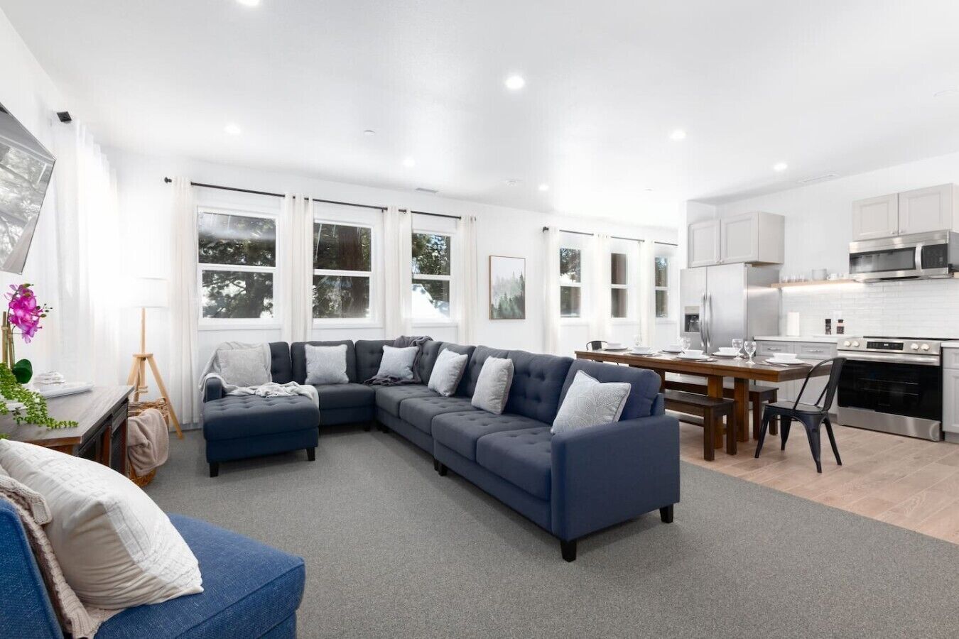 A modern living room with a sectional sofa, a dining table, and a kitchen area with white cabinets and stainless steel appliances.