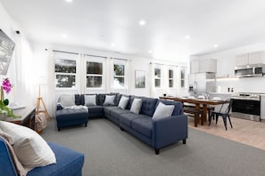 A modern living room with a sectional sofa, a dining table, and a kitchen area with white cabinets and stainless steel appliances.