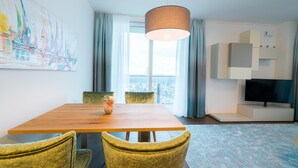 Una sala de estar moderna con una mesa de comedor de madera, sillas tapizadas en verde terciopelo, un cuadro abstracto grande, un televisor y vistas de la ciudad a través del ventanal.