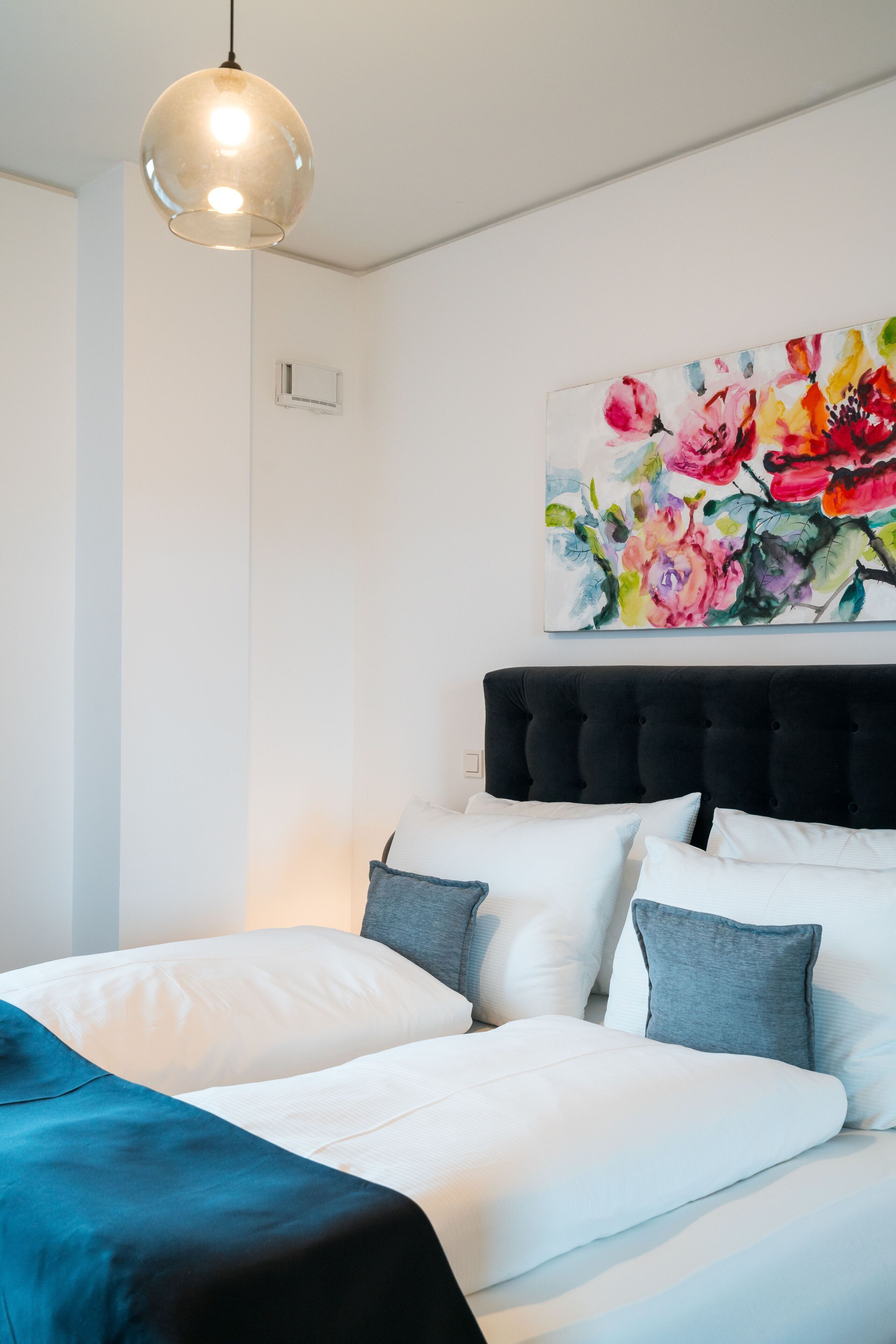 A neatly made bed with white and gray bedding, a black headboard, and a colorful floral painting above it.