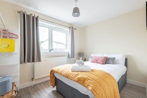 Een slaapkamer met een bed, een raam, een radiator, een hanglamp en een gele tas op een plank.