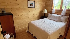 Een slaapkamer in een houten hutje, met een bed, een nachtkastje, een ingelijste foto aan de muur en een raam met gordijnen.