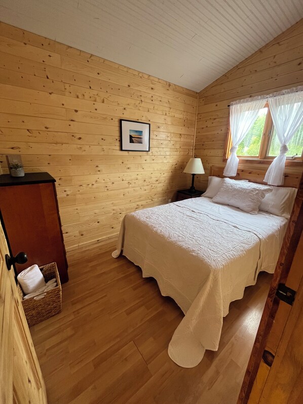A wooden cabin bedroom with a bed, a nightstand, a framed picture on the wall, and a window with curtains.