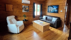 Een gezellig interieur van een blokhut met houten wanden, een plafondventilator, een bank, een witte fauteuil, een salontafel en een tv aan de muur.