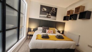A modern bedroom with a large bed, a headboard, bedside lamps, a window, and a wall-mounted shelf.
