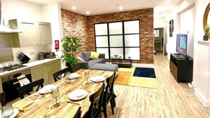A modern kitchen with a dining area, a living room with a sofa and a TV, and a large window with blinds.