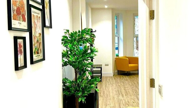 A hallway with framed botanical prints on the wall, a potted plant, and a modern orange chair in a bright room with wooden flooring.