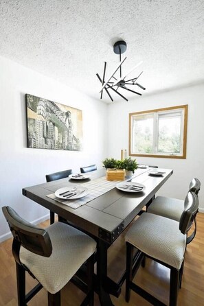 En matplats med ett långt bord, stolar, en modern taklampa, en målning som föreställer en stadsbild och ett fönster med utsikt.
