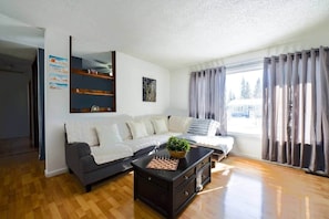 Una sala de estar con un sofá, una mesa de centro con un juego de ajedrez, una ventana con cortinas y una pared con un póster.