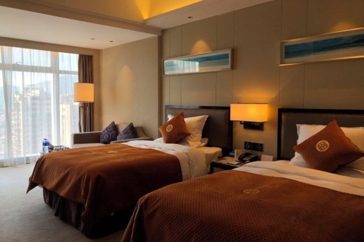 Una habitación de hotel con dos camas, un ventanal con cortinas, un cuadro colgado en la pared y una mesita de noche con lámpara.