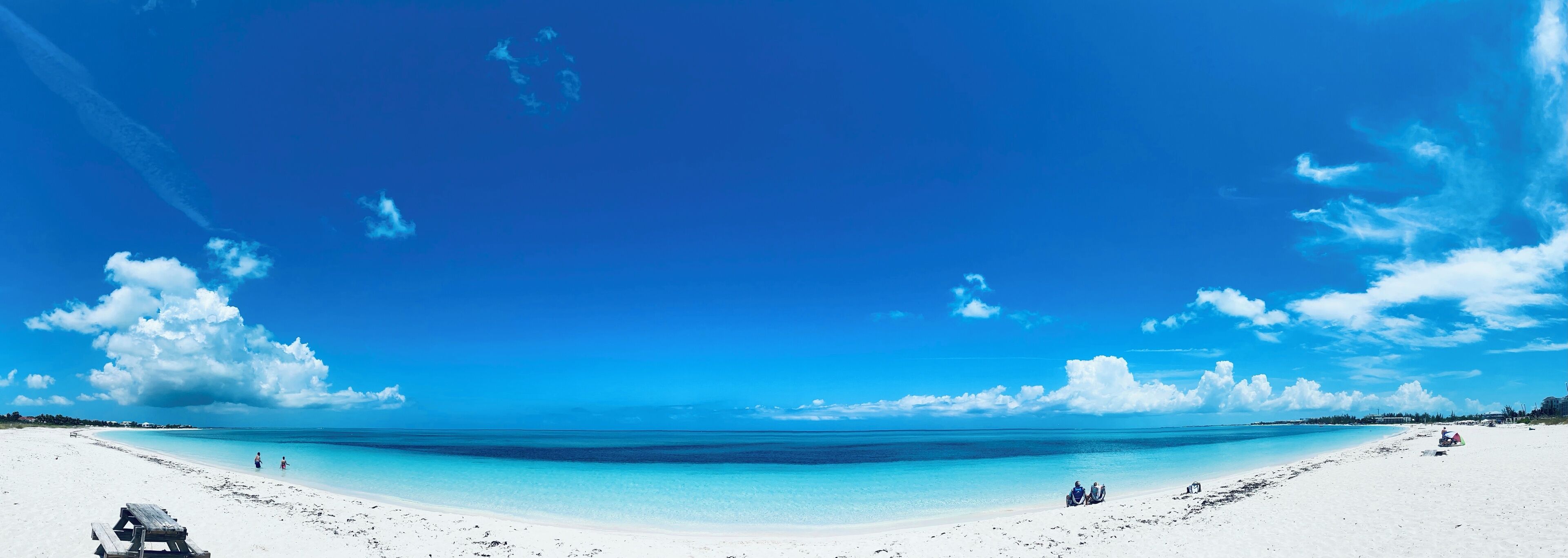 Ein weiter, sandiger Strand mit klarem, türkisblauem Wasser, einigen Personen und einer Holzbank.
