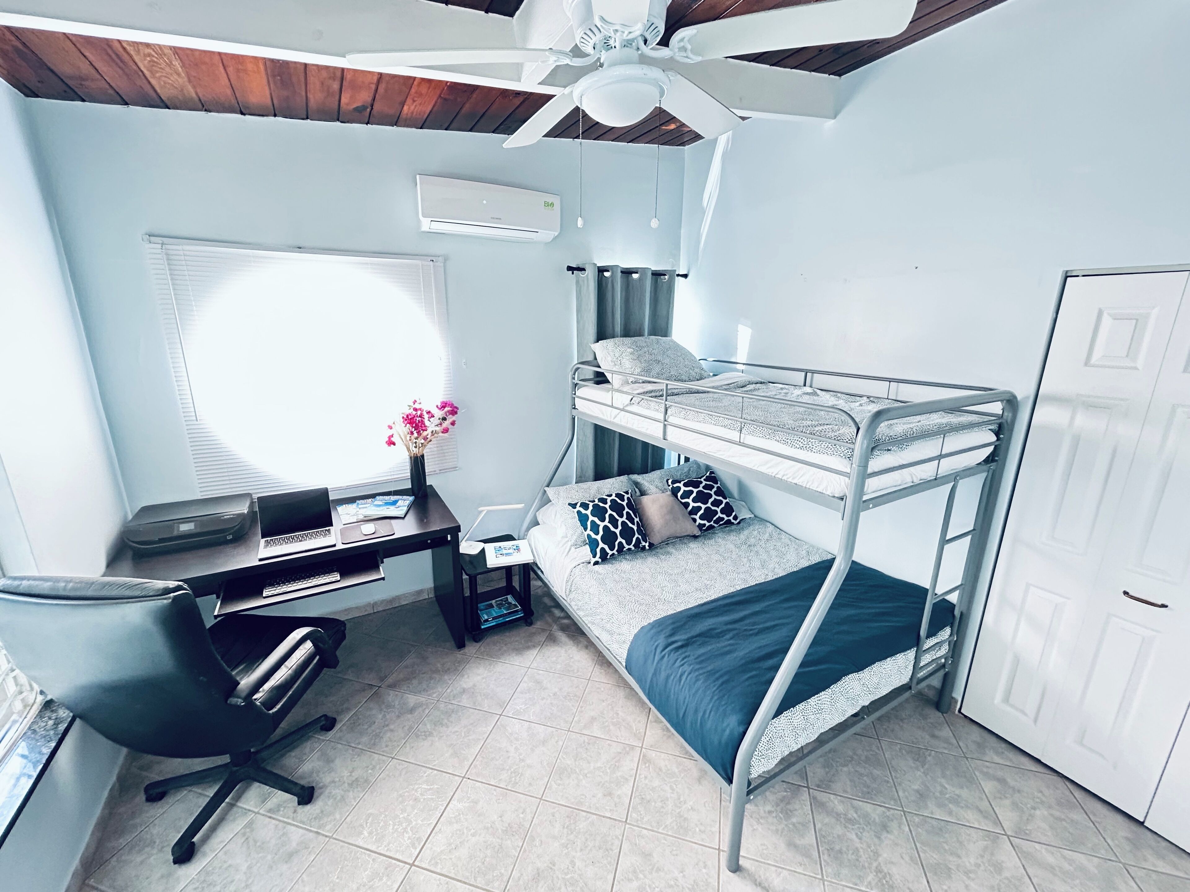 Une petite pièce comprenant un lit superposé, un bureau avec un ordinateur, une chaise et un ventilateur de plafond.