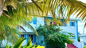 A building with a white exterior and blue accents, a red staircase, and lush greenery including palm trees and flowering plants. - Blue Bird Villa, Open & Breezy Island Home...minutes from the beach! (Providenciales)