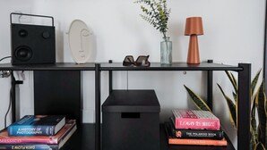 A black desk with a speaker, a vase with a plant, a pair of sunglasses, and a lamp. There are books stacked on the desk and a black speaker on the floor.