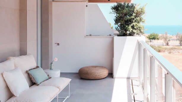 A balcony with a white sofa, a round ottoman, and a potted plant.