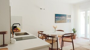 A modern kitchen with a white countertop, a sink, and a fruit basket. There is a dining area with a round table and wooden chairs.