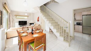 Una cocina moderna con un refrigerador de acero inoxidable, un comedor con mesa y sillas de madera, y una escalera con barandilla de vidrio.