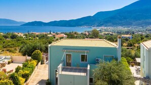 Una zona residencial costera con un edificio turquesa de teja, rodeado de vegetación y con vistas a una masa de agua y montañas a lo lejos.