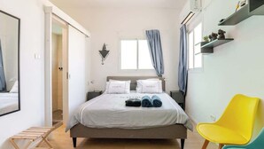 Un dormitorio con cama, un tendedero de madera, una silla amarilla y una ventana con cortinas.
