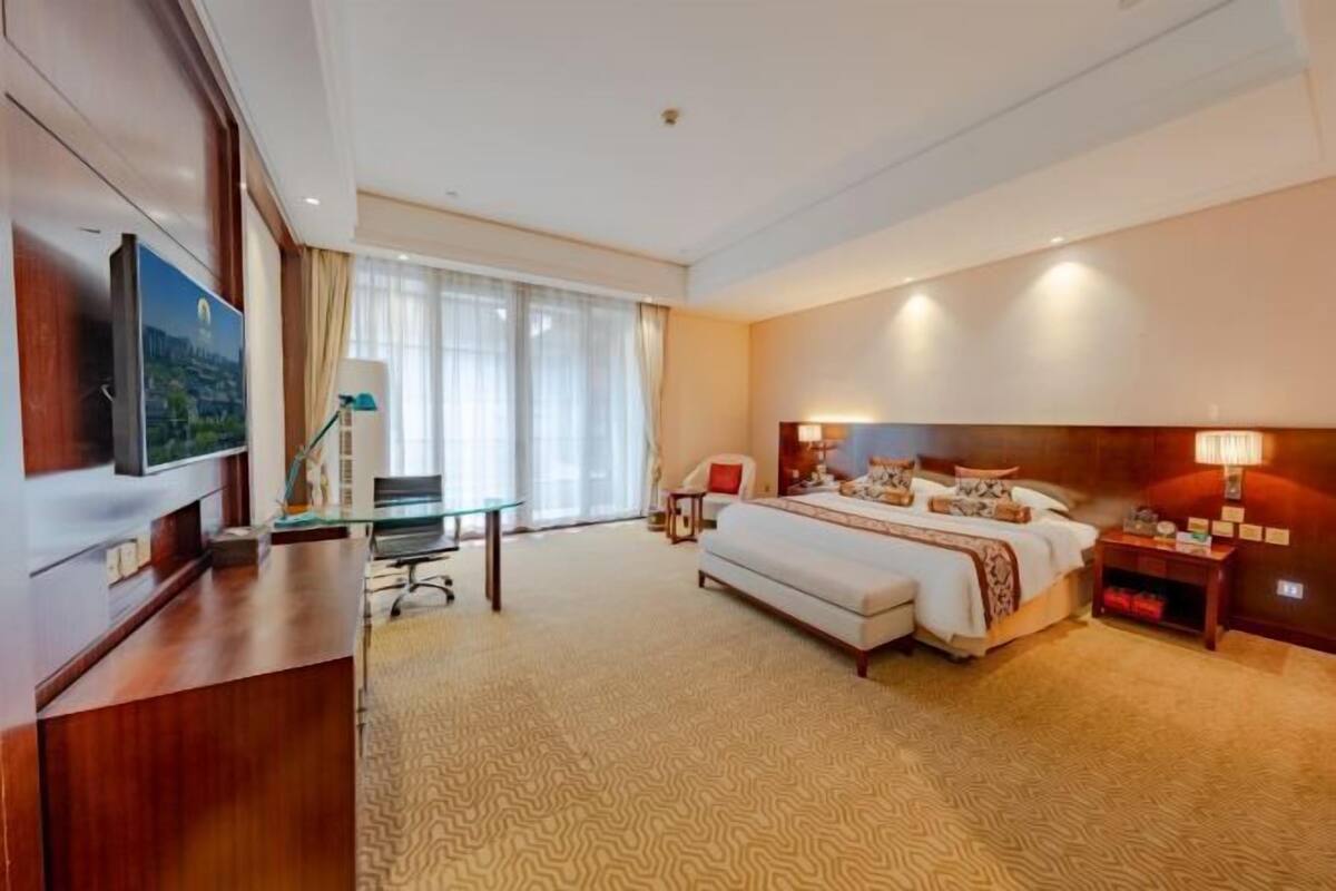 Quarto de hotel com cama, escrivaninha, cadeira, televisão e uma janela grande com cortinas.