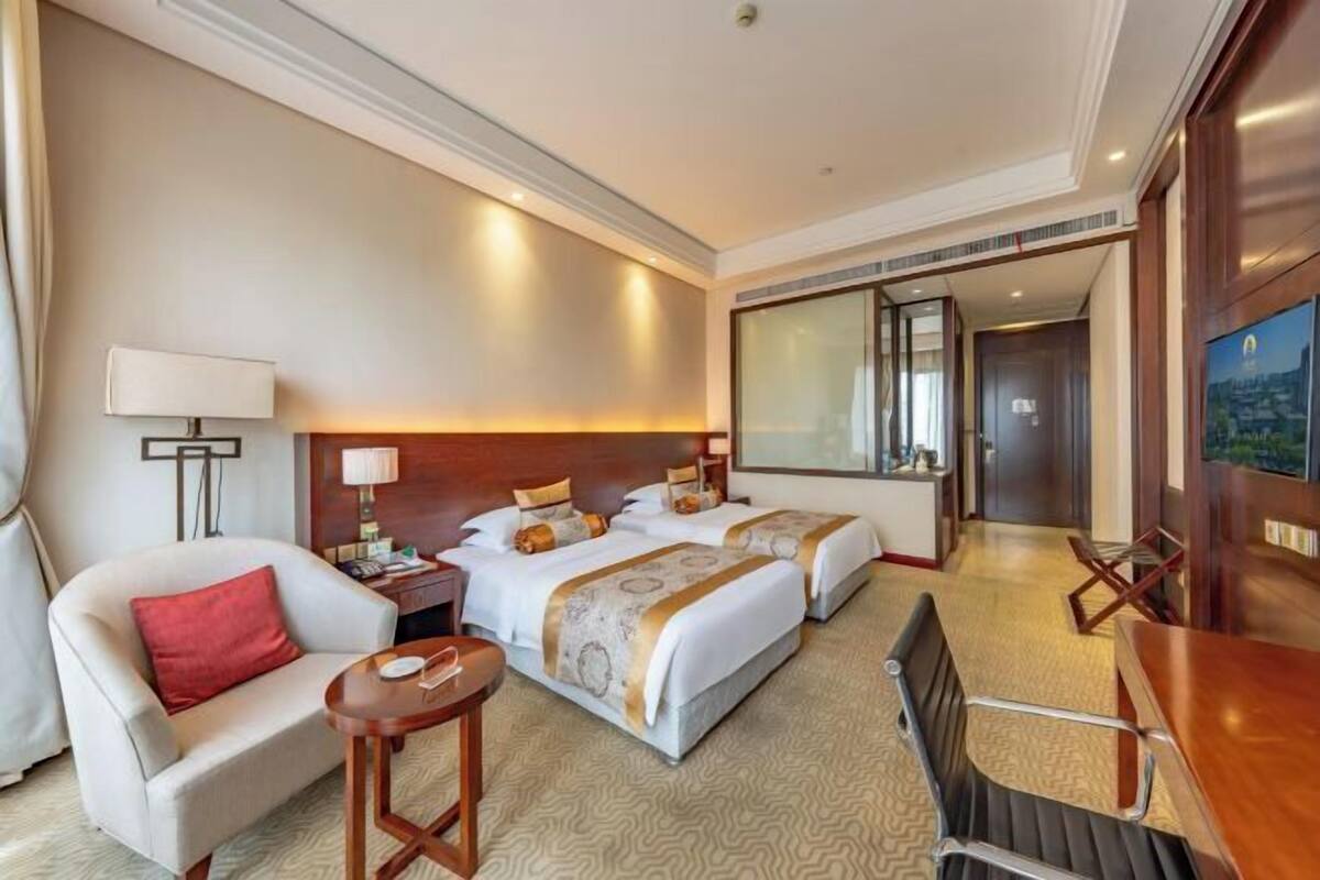 Quarto de hotel com duas camas, uma escrivaninha, uma cadeira, um abajur e um espelho grande.